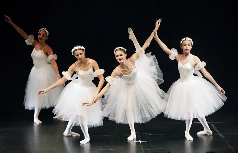 Four teenage ballerinas in long white tulle skirts and flower crowns, one lifting a leg high