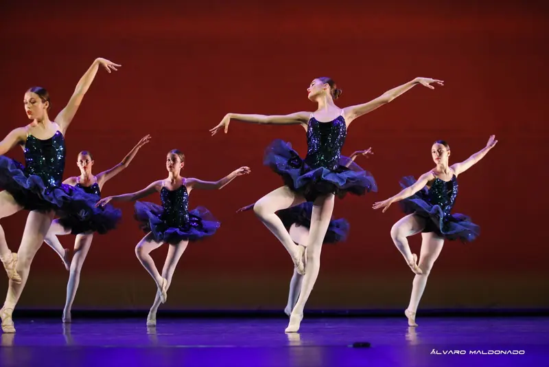 Five ballerinas in navy sequined tutus balance on one leg against a deep red backdrop