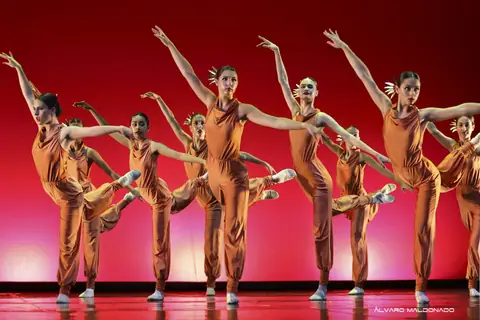 Dancers in orange jumpsuits stretch their arms and legs in unison against a vivid red backdrop